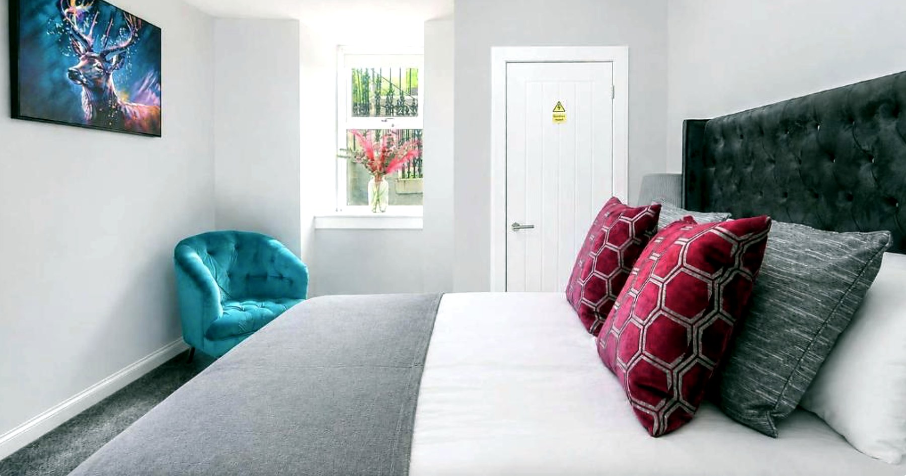 Double bedroom with a grey buttoned headboard, red cushions and a teal armchair by the window
