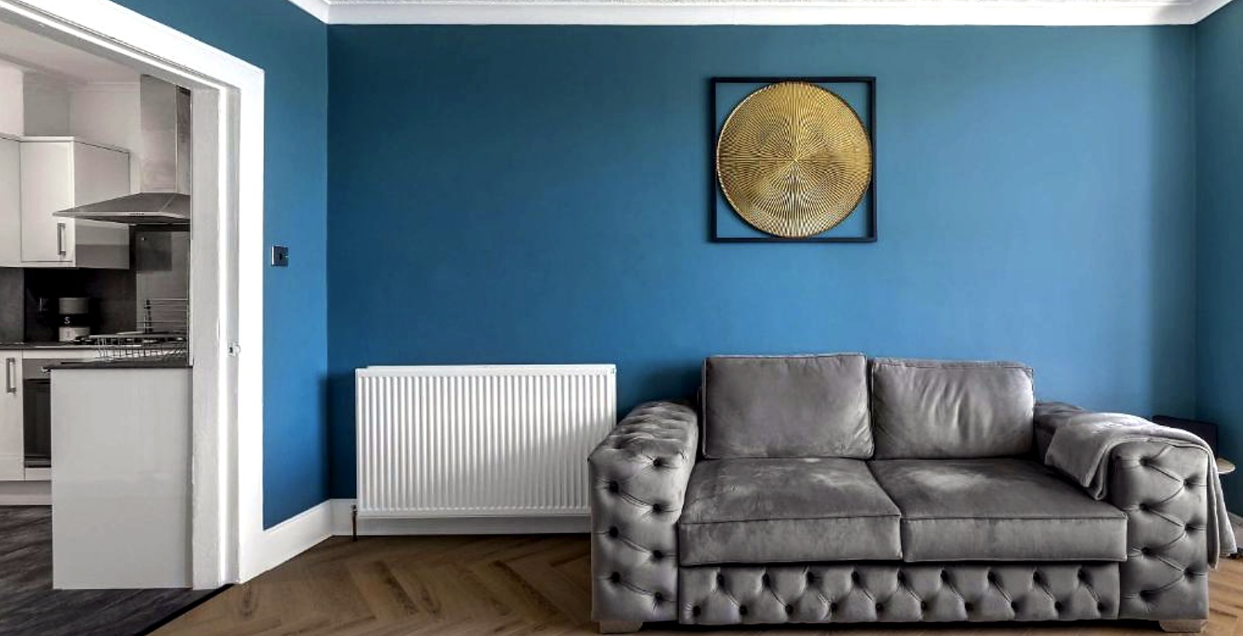Grey buttoned sofa against a deep blue wall with the kitchen through an open doorway