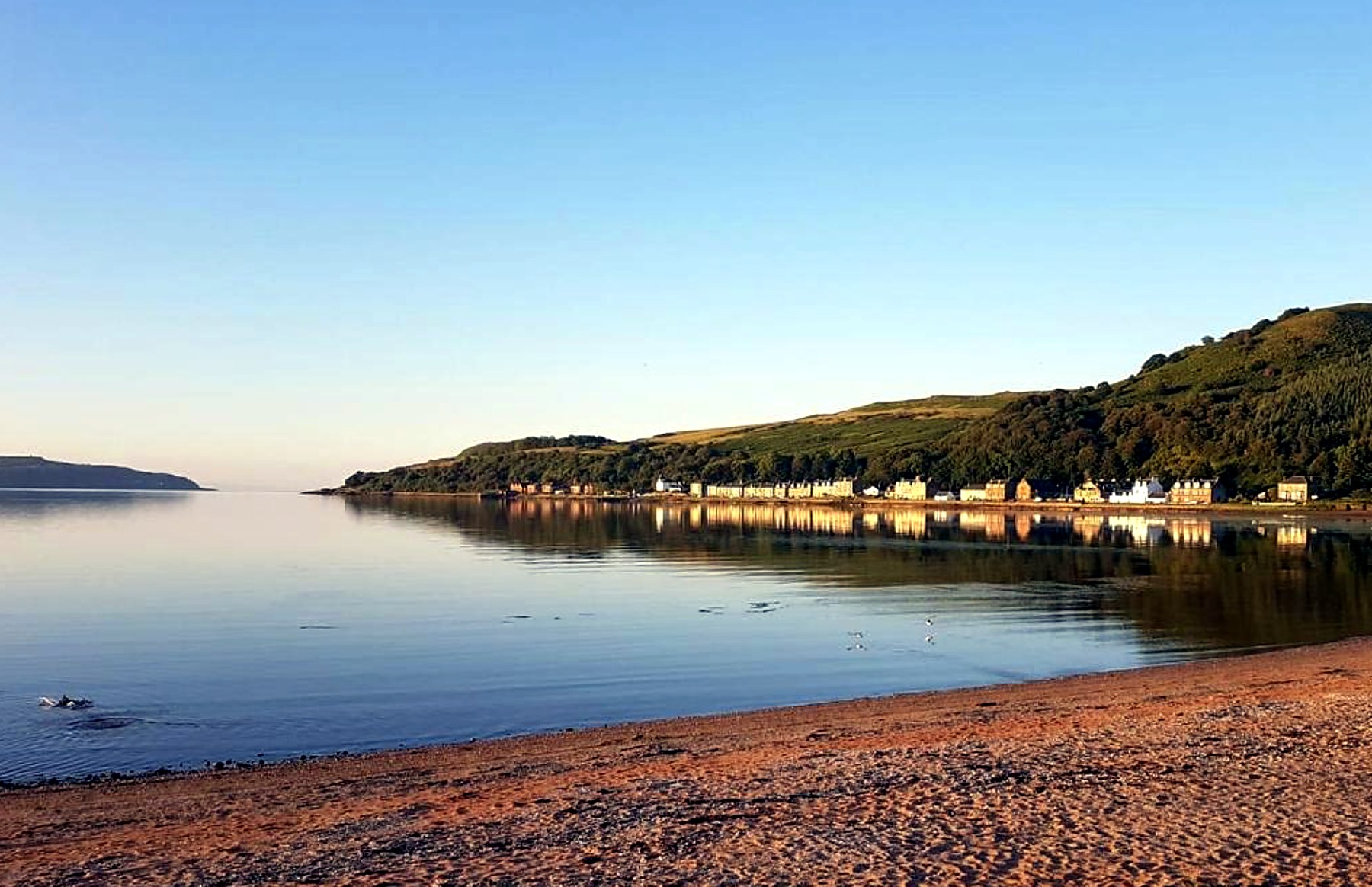 Still morning water and sand at Kilchattan Bay with the village along the shore