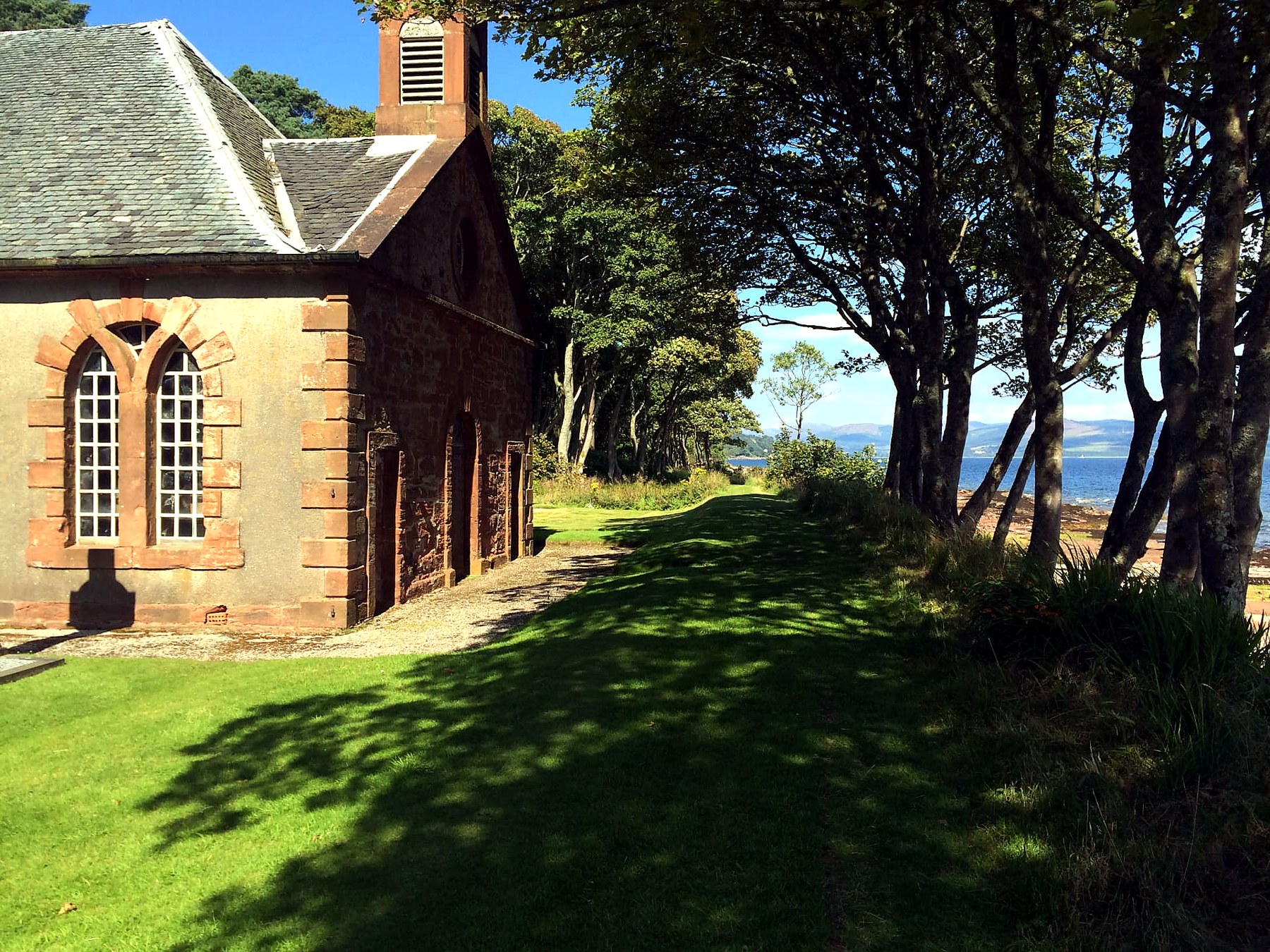 A small harled chapel beside the shore path on the Isle of Bute, with the mainland hills across the water