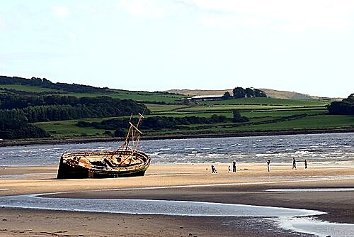 The old wooden shipwreck resting on the sand at Ettrick Bay, Isle of Bute, with visitors walking the beach and green hills behind