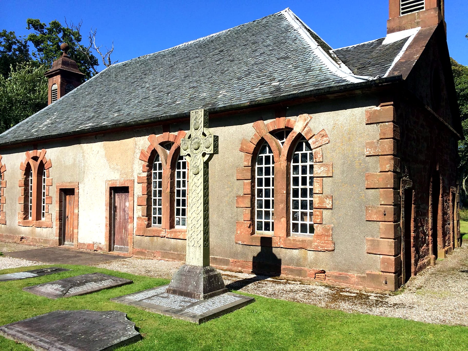 A carved Celtic cross and old gravestones beside a red sandstone chapel on the Isle of Bute