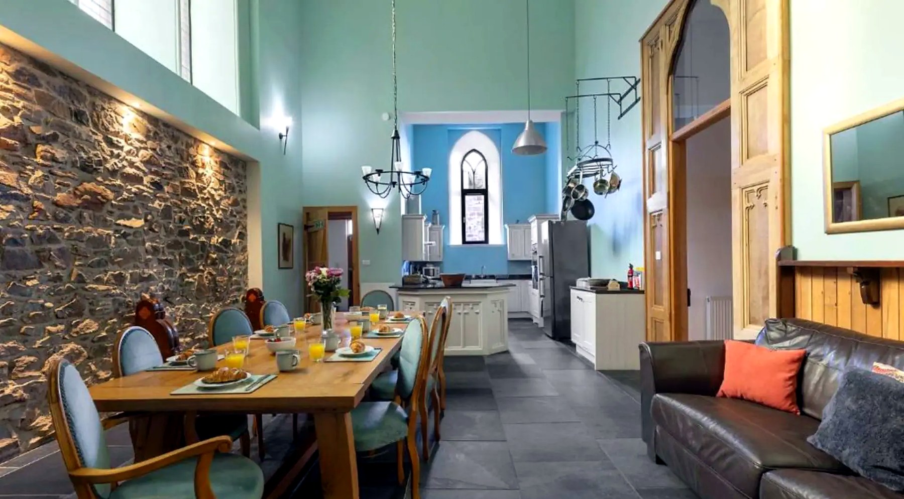Double-height dining hall with long oak table, exposed stone wall and kitchen beyond in the converted church