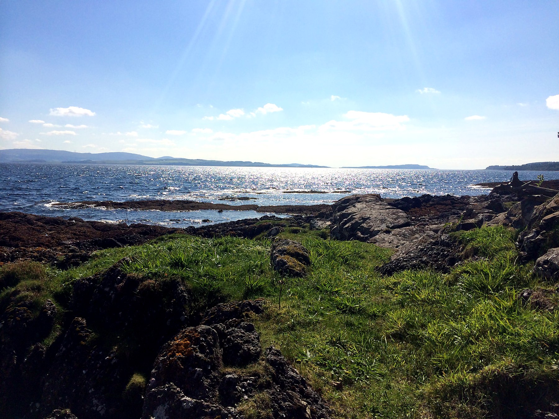 Grass and volcanic rock shoreline with sunlight sparkling on the Firth of Clyde, Isle of Bute